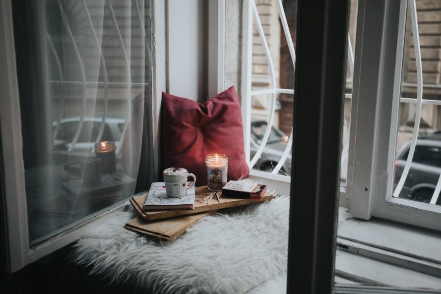 beside an open window is a maroon pillow and a tray with a candle, mug, and books