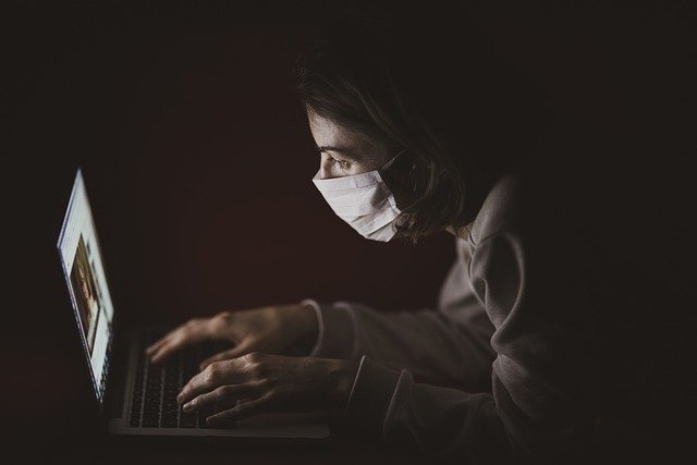 a person wearing a mask types on a laptop in a dark room