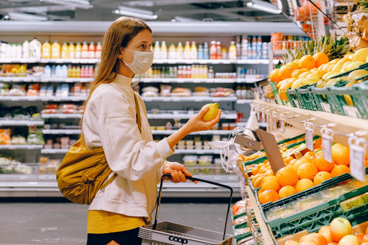 A woman is grocery shopping. She wears a yellow t-shirt and beige jacket and a face mask. She is holding a citrus fruit.