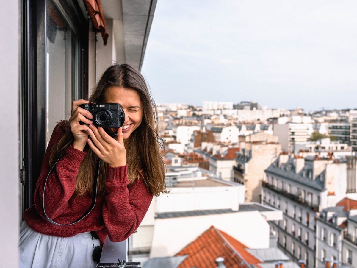 A woman wearing a maroon sweater on a balcony holds a camera. The city is visible behind her.